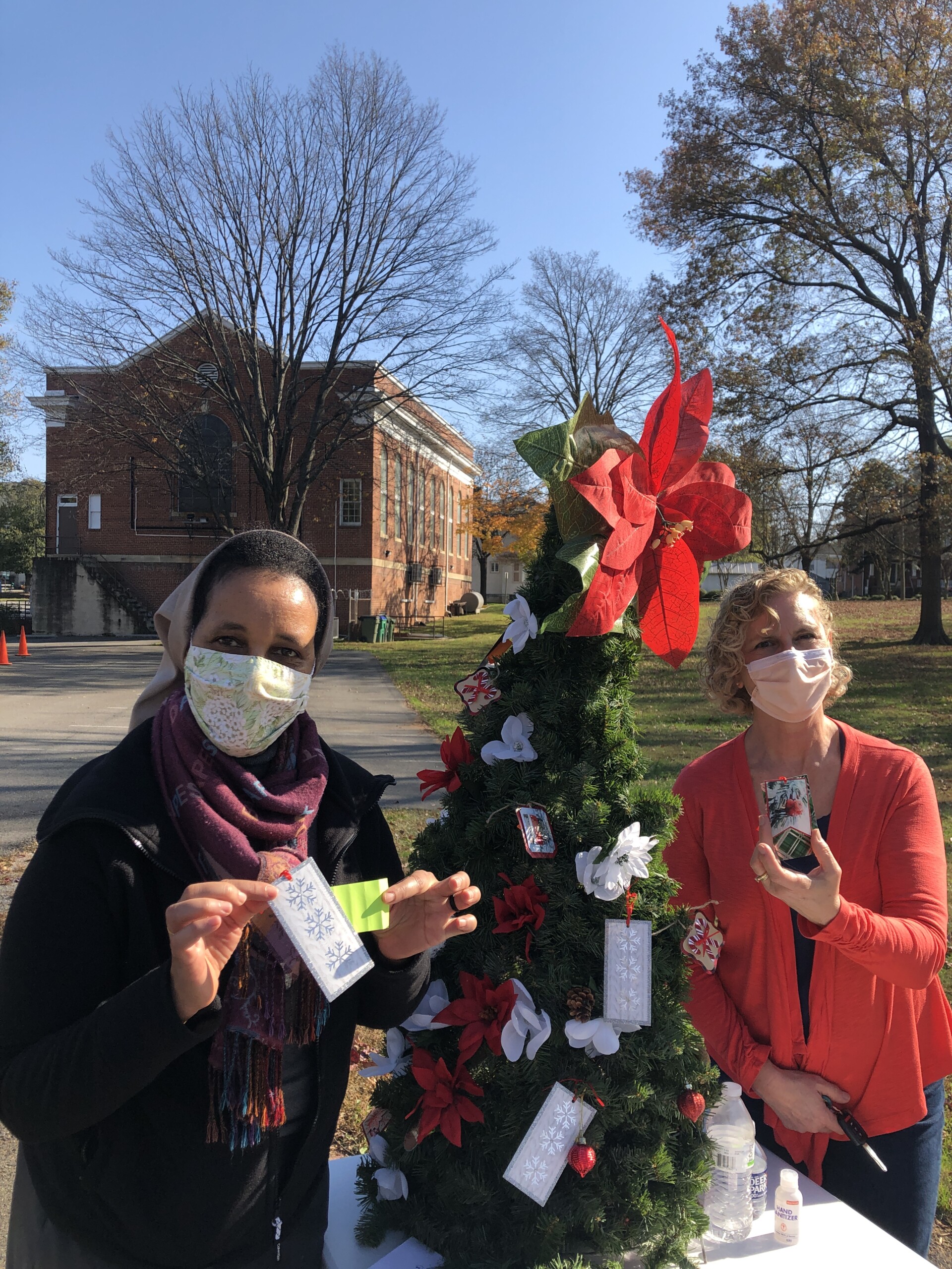 Two women, both wearing masks, in front of a small tree decorated with gift tags and a red bow